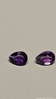 Two pear shaped deep purple amethyst gemstones placed side by side on a neutral surface with clear lighting to show cut and color.