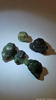 Five natural rough emerald crystals displayed on a light surface, varying green hues and matrix presence visible