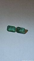 Two natural emeralds placed side by side on a light surface, showing their rectangular cut and green color with visible inclusions.