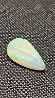 Close-up photo of a pear-shaped natural Ethiopian opal cabochon showing vibrant rainbow flash on black textured background.
