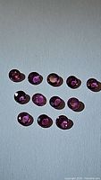 Photo showing 13 natural round cut rubies in pink, purple, and red hues arranged on a light surface.