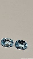 Pair of natural oval cut sky blue topaz gemstones displayed on neutral background, showing their clarity and faceting.