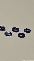 Photo showing five oval cut blue-purple tanzanite gemstones on a beige background.