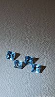 Four natural Swiss blue topaz princess cut gemstones arranged in a row on a textured surface, showing color, cut and clarity.