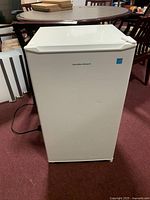 Front view of white Hamilton Beach mini fridge closed, placed on red carpet flooring with dining table in background.