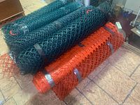Photo showing multiple rolls of green and red plastic mesh snow fencing on a tiled floor indoors, some secured with tape, varying in length and diameter.