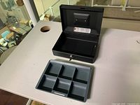 Open steel cashbox showing black interior and removed blue-gray plastic compartmentalized tray on table.