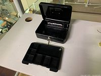 Steel cashbox open with removable cash tray separated, showing interior compartments and key inserted in lock.