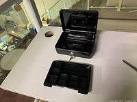 Black steel cashbox open showing removable tray with multiple compartments. Front lock with key inserted visible.