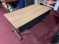 Full view of rectangular wooden laminate top table with silver metal legs and caster wheels on carpet floor.
