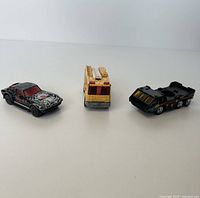 Front angled view of three Matchbox cars showing black/red spider web car, yellow/red emergency vehicle, and black six-wheeled vehicle.