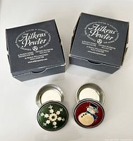 Two enamelled pewter memory boxes with snowflake and snowman designs shown next to their original packaging boxes from Aitkens Pewter.