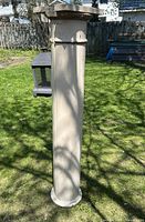 Photo showing full length of the pillar standing upright on grass, with a bird feeder and fence background. Shows smooth cylindrical beige pillar with decorative molding.