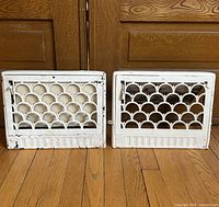Front view of two white metal antique vent covers with scalloped decorative cutouts, placed on wooden floor against cabinet doors.