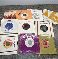 Photo showing nine 45 vinyl records with various labeled sleeves in varying condition.