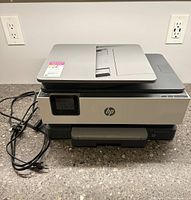 Front view of HP OfficeJet Pro 8034e printer with power cord coiled beside it on countertop, printer lid closed.
