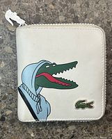 Front view of white Lacoste wallet with cartoon crocodile graphic and Lacoste logo patch, zipper fully closed.
