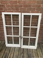 Two white-painted wooden century window frames with six glass panes each, leaning against a brick wall, showing condition and metal latches.