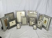 Photo showing 12 assorted silver picture frames of various sizes and designs arranged on a white fabric background.