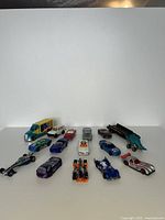 Front view of 15 vintage Hot Wheels die-cast vehicles arranged on a white board background showing various models including a dinosaur-themed truck, race cars, and sports cars.
