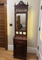 Full view of the tall wooden hall stand showing carved top, mirror, tiles, drawer and cabinet door.