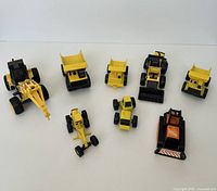 Eight vintage Tonka trucks arranged on white background, showing variety of construction equipment including dump trucks, bulldozers, and loaders in yellow, black, and orange.