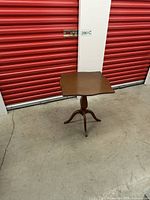 Full view of antique side table showing the shaped rectangular top and three-legged pedestal base against a red storage door background.