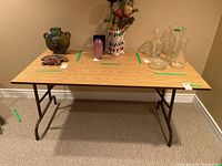 Front view of a rectangular folding table with wood veneer top, supported by black metal folding legs, placed on carpeted floor against a wall.