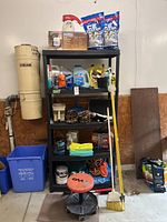 Full lot view on black shelving unit showing various garage and cleaning supplies, bags of sidewalk salt, bottles, stool, broom, cords and more.