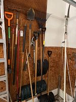Garage corner with various garden tools hanging on pegboard including shovels, rakes, and plastic snow shovels stacked below.