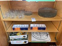 Shelves with 12 clear glass cups and 5 saucers on top shelf, stacked glass plates on right, two boxes labeled Finland Iittala Design Tapio Wirkkala containing glasses, and boxed Arcoroc glassware and a round divided glass serving platter.