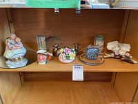 Shelf displaying various porcelain and brass collectibles including a floral porcelain arrangement, brass duck with umbrella, brass candle holder, Stafford China figure, Lancaster Uncle Tom Toby jug, and porcelain bird figures.