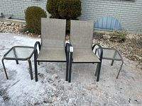 Two stacking patio chairs with grey mesh fabric seats and backs flanked by two square tempered glass top side tables on frosty brick patio surface.