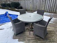 View of outdoor patio set on wooden deck with snow around: round tempered glass table, four grey faux wicker chairs arranged around it.