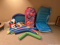 Overview showing two boogie boards, multiple pool noodles, inflatable lounge float, colorful towels, orange bucket with pool accessories, and a football.