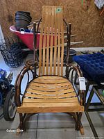 Front view of twig and bent wood rocking chair with slatted wood seat and backrest. Curved armrests made from natural wood branches. Blue cushion visible nearby but separate.