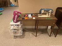 Wide shot showing Reynolds sewing machine placed on a brown wooden sewing table with hinged top. Also visible is a plastic utility drawer unit and various sewing accessories on the table surface.