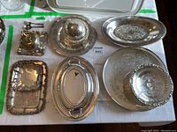 Overview of seven silver plated serving pieces including trays, butter dish with dome, covered veggie server, pedestal cake plate, and more arranged on white cloth.