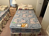 Front view of the single mattress with matching floral bed skirt and folded linens on top. Shows mattress top and side pattern as blue quilted with geometric patches.