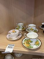 Four tea cups and saucers displayed on a shelf, showing three matching Royal Albert yellow rose sets and one Paragon set with a pink/yellow rose design.