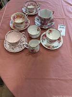 Photo shows 5 cups and matching saucers, a creamer and sugar bowl on a pink tablecloth. Various floral and pink patterns with gold trim. Visible chips on pieces.