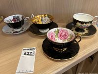 Four vintage teacups and saucers displayed on a wooden shelf showing floral and black and gold designs