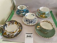 Five vintage teacups and saucers arranged for display, showing variety of designs and colors including turquoise, green, blue, white, and gold trim.