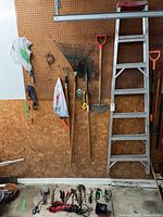 Wall with aluminum step ladder, garden rake, shovel, shovel edger, and other hanging garden tools with various hand tools and power tools on floor.
