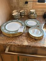 Stack of dinner plates, luncheon plates, bread plates, cups and saucers arranged on a countertop showing Asia pattern with green and gold borders.