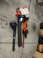Full view of the Black & Decker Vortex electric blower with nozzle attachment and battery powered hedge trimmer with charger on garage floor.