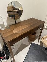 Brown wooden vanity desk with two drawers and round mirror on black metal stand, shown in room setting.