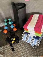 Rolled black thick foam exercise mat leaning against wall with stacked neoprene dumbbells, TRX straps on floor, and folded colorful towels beside them.