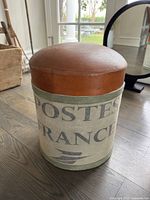 Oblique side view of round stool with brown leather top and fabric base with French postal print.