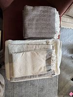 Five assorted blankets and throws stacked showing different woven textures and neutral colors, folded on a chair and floor.
